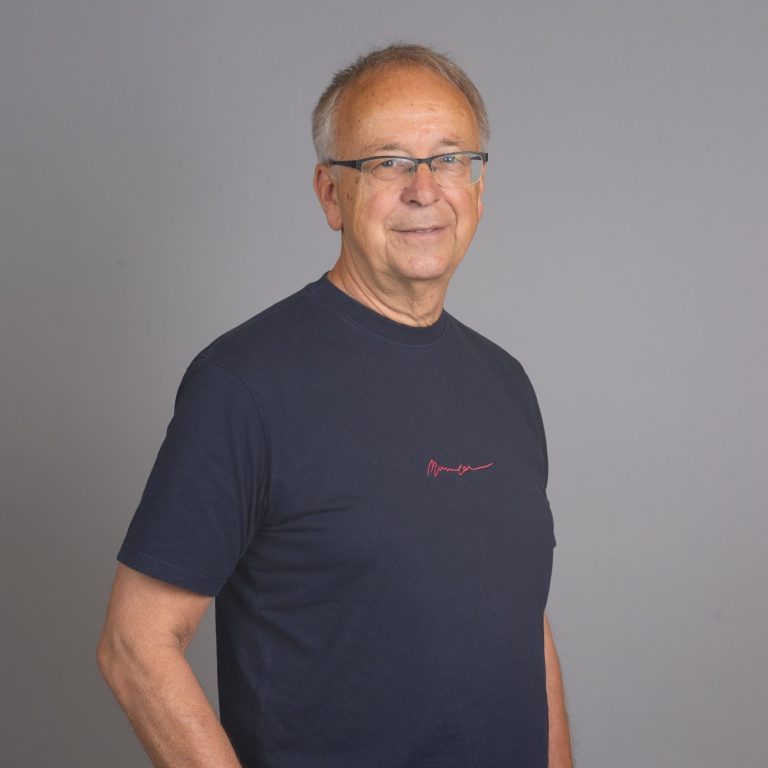 Älterer Mann mit Brille in schwarzem T-Shirt, neutraler Hintergrund.