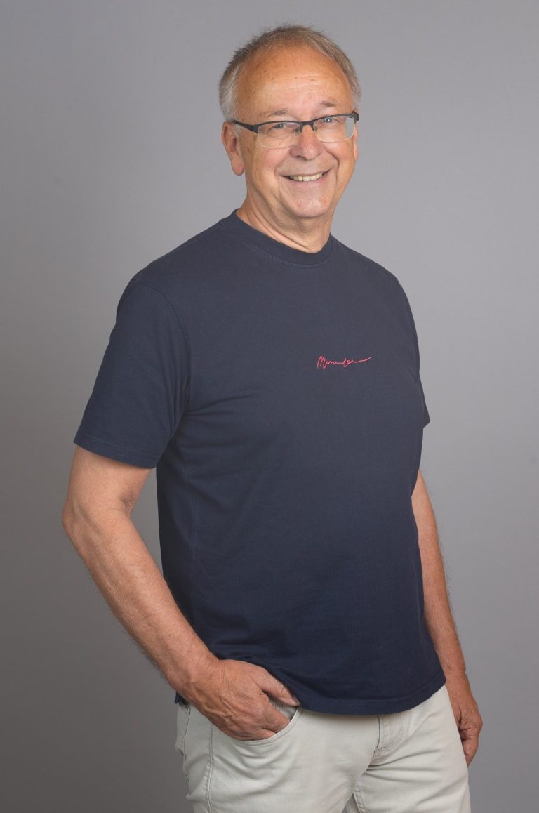 Andreas Bieler - Feng Shui und Geistiges Heilen Lächelnder Mann in einem dunkelblauen T-Shirt, mit Brille, im Stehen vor neutralem Hintergrund.