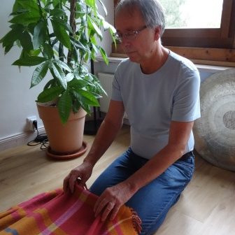 Seelenzaubern in Aktion Eine Person sitzt auf dem Boden und faltet eine bunte Decke neben einer Pflanze.