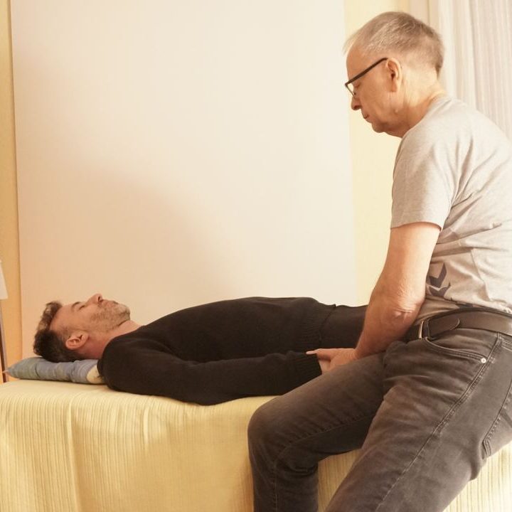 Die geistige Wirbelsäulenaufrichtung in München Zwei Männer in einer Behandlungssituation, einer liegt auf einer Liege, der andere sitzt daneben.