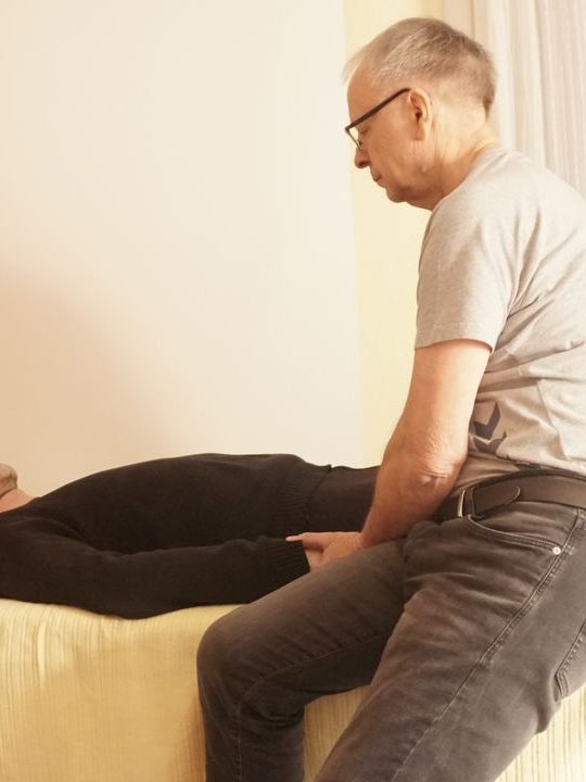 Einzelbehandlung geistiges Heilen Ein Mann sitzt auf einem Bett und unterstützt eine liegende Person beim Entspannen.