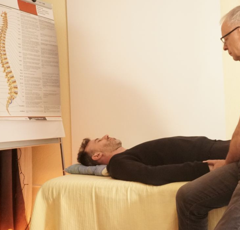 Ein Mann liegt auf einer Liege, während ein anderer ihn beobachtet und eine Wirbelsäulenkarte zeigt.