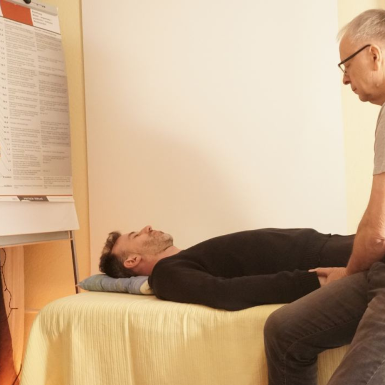 Die energetische Wirbelsäulenaufrichtung in München Mann liegt auf einer Liege, während ein Therapeut ihn behandelt. Hintergrund mit Poster.