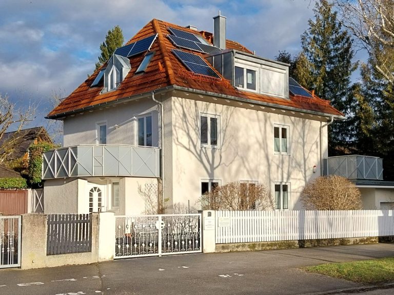 Feng Shui Beratung und Praxis Homöocampus in München Helles Haus mit rotem Dach, Solarpanelen und Gartenzaun, umgeben von Bäumen.