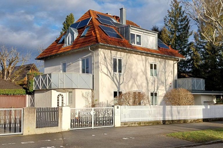 Feng Shui Berater in München Helles Haus mit rotem Satteldach, Balkon und Solarpanels, umgeben von einem weißen Zaun.