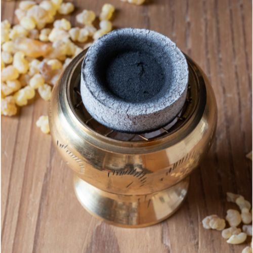 Ein runder, grauer Kohletablette auf einem goldenen Räuchergefäß, umgeben von Harzkörnern.