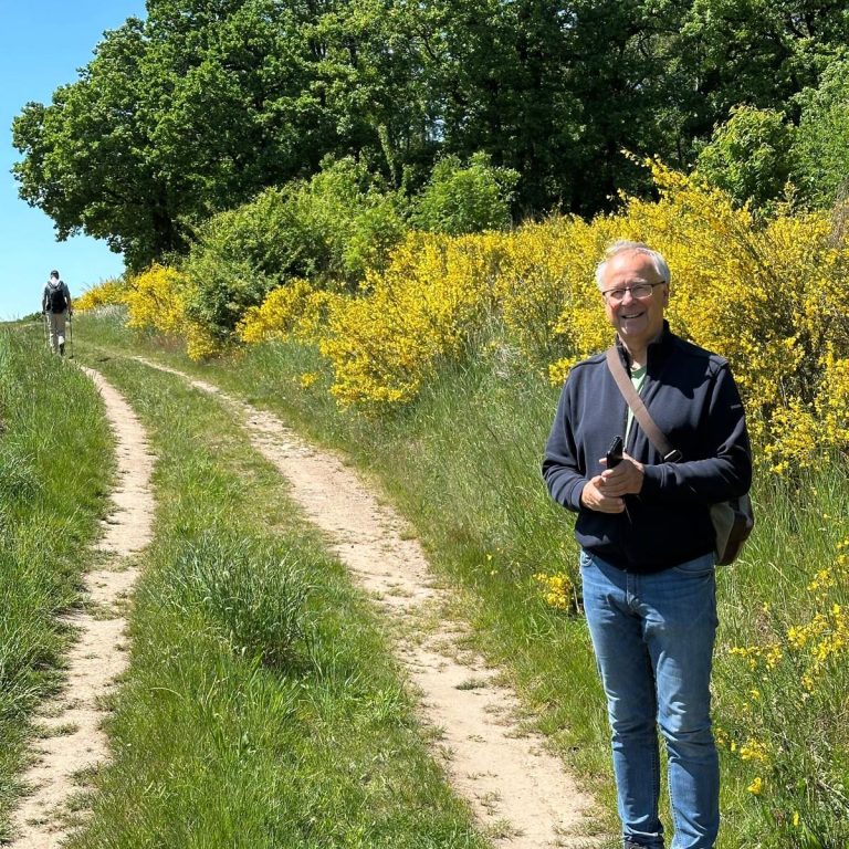 Mann steht auf Weg neben blühenden Sträuchern, eine andere Person im Hintergrund.