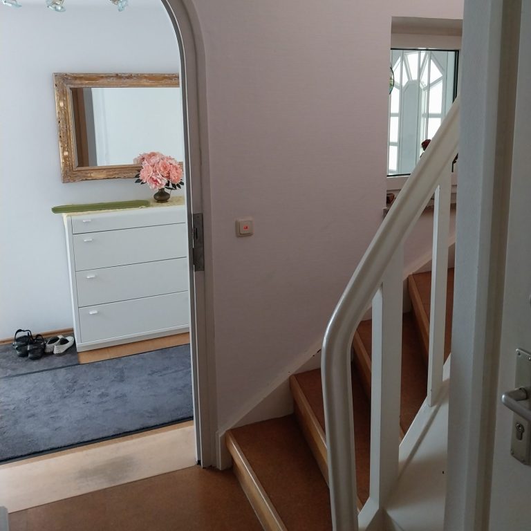 Innenansicht eines Hauses mit Treppe, Kommode und Spiegel im Eingangsbereich.